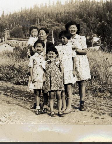 Black-and-white image of women and children standing on a gravel road in front of a field. Partially obscured.