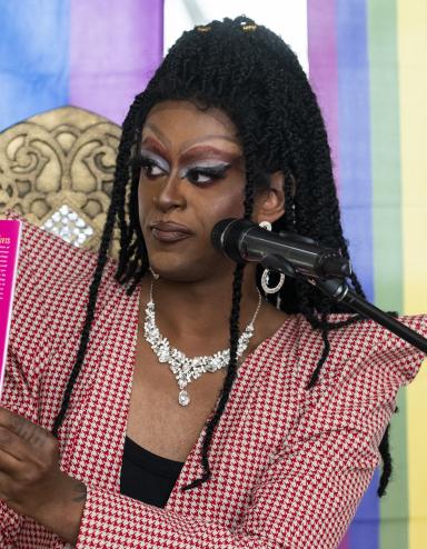 A drag queen in heavy makeup and a wig of long tiny braids holds up a children’s book to show an audience. The backdrop behind her is a pastel rainbow. Partially obscured.
