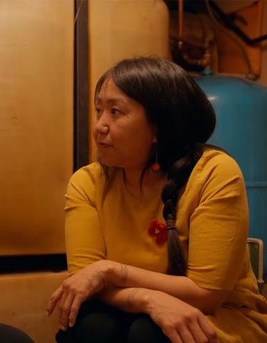 Deux femmes inuites sont assises dans une pièce sombre, l’une en face de l’autre, et discutent de manière intime. L'une porte un chandail rouge, l'autre un chandail jaune. Partially obscured.