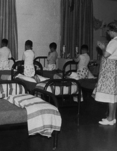 A group of boys in pyjamas kneel on single beds, heads bowed and hands clasped as if in prayer. A woman stands in the room, her hands clasped in the same way. Partially obscured.