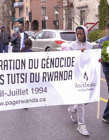 A group of people walking down a street with sign that reads “Commémoration du génocide contre les Tutsi du Rwanda. Avril-Juillet 1994. www.pagerwanda.ca”. Partially obscured.