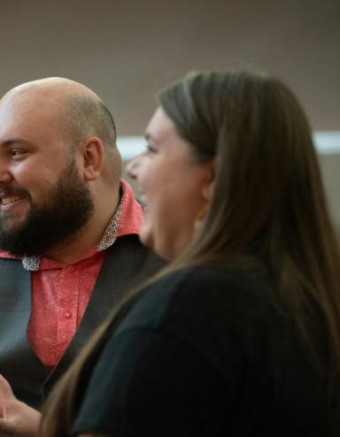 A man and woman sit together at a dinner table, smiling warmly as they listen to other guests. The man wears a salmon shirt with a gray vest, and there are small candles and drinks on the table, creating a cozy intimate atmosphere. Partially obscured.