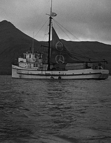 A boat in the water with mountains and clouds in the background. Its sail is decorated with large peace signs. Partially obscured.
