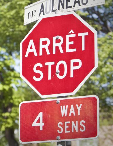 A stop sign and street sign in both English and French are seen in front of trees. Partially obscured.