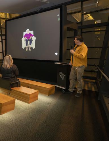 A small group of people sitting on wooden benches in a theatre face a screen showing an older woman seated in a chair. A man in a yellow shirt is standing to the right of the screen holding a microphone. Partially obscured.