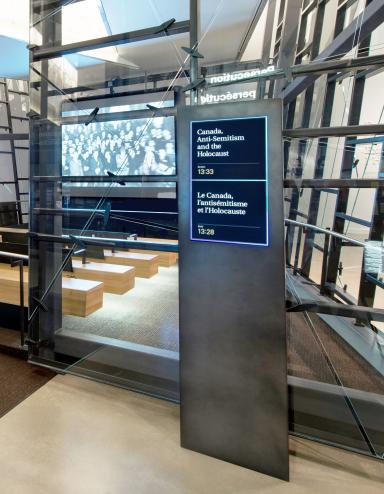 Uneven glass walls surround a theatre with a screen and 4 benches facing it. Outside the theatre entrance is a sign indicating the film selected to play. Partially obscured.