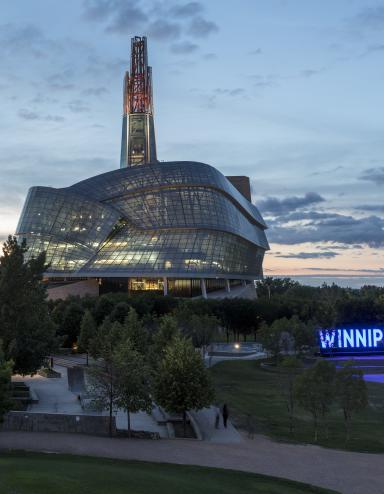 Exterior photo of the Canadian Museum for Human Rights’ glass windows and tower, taken against a prairie sky at dusk. The Winnipeg sign at the Forks is situated on the right side of the image. Multiple office towers and downtown lights are on the left-hand side of image. Partially obscured.