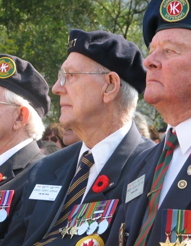 Three older men in Canadian Legion uniforms sit beside each other. They are all wearing Remembrance Day poppies on their uniforms.