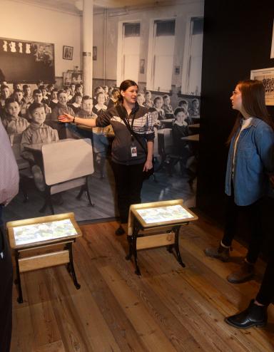 A visitor looks at a gallery niche on residential schools at the Canadian Museum for Human Rights. Two small desks sit in the middle of the niche. A large image of Indigenous students sitting at desks at a residential school is on the back wall of the niche, with images and artifacts on the side walls.