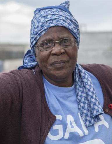 A head-and-shoulders portrait of Gogo Gladys Tyophol. She is wearing glasses and a blue patterned kerchief wrapped around her head. Her shirt says GAPA and a red AIDS ribbon is attached to her sweater.