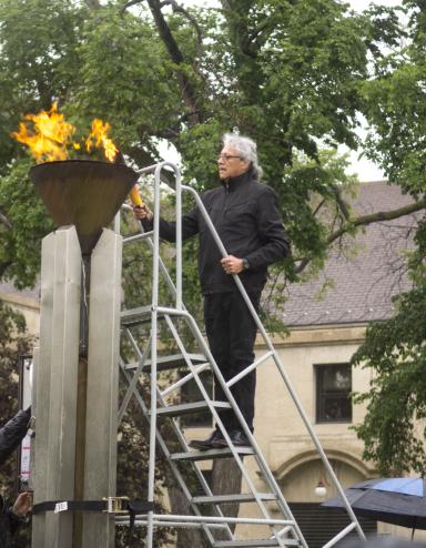 A man carrying a torch stands on a ladder to light a flame within a copper cauldron.