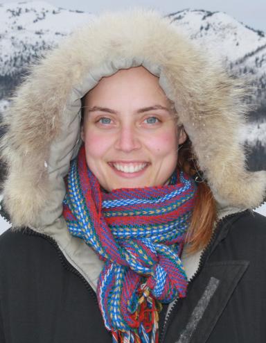 Smiling female wearing a fur-lined parka posing in front of a mountainous terrain.