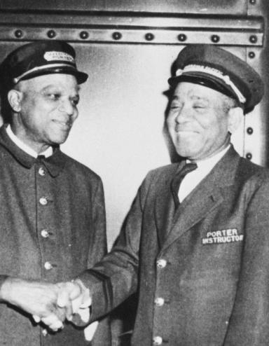 A black and white photo of four men in train porter uniforms. All of the men are smiling, and the two men in the middle appear to be shaking hands.
