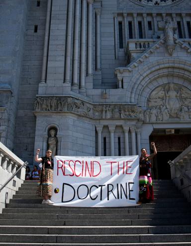Two people in braids and ribbon skirts raise fists and hold a large cloth banner reading “RESCIND THE DOCTRINE” on the steps of an enormous cathedral.