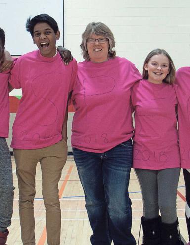 Six teenagers and a middle-aged woman stand with their arms around each other and are smiling for the camera. They are all wearing pink shirts.