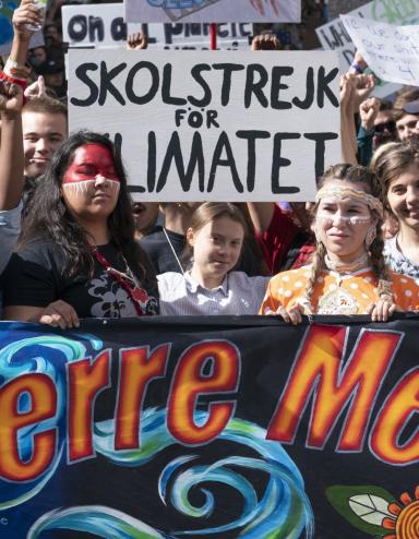 A crowd of youth hold protest signs and stand behind a large banner that reads “La Terre Mère,” or “mother earth” in English.