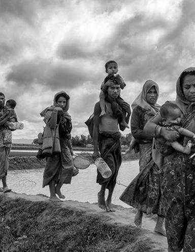 A group of Rohingya women and men carry young children and belongings as they walk in a line over an earthen dike over a stretch of water.