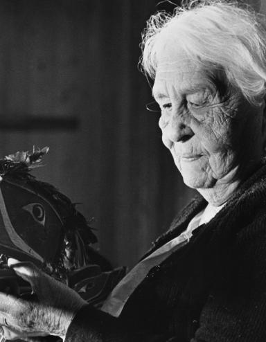 A black and white image of an elderly woman with white hair holding a mask that is shaped and painted to represent a bird’s head. It features a long beak with an open mouth and feathers attached to the top of the head. The woman is looking directly at the mask she is holding.