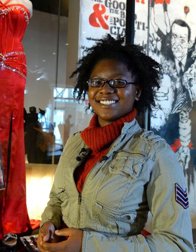 A smiling woman stands in front of a red prom dress and a black tuxedo mounted on mannequins. Both the dress and suit are on display behind a glass case.