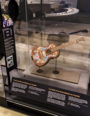 A row of museum display cases with various artifacts inside. A guitar is displayed under glass. The guitar has a colourful floral print with a bird hand-painted onto its front.