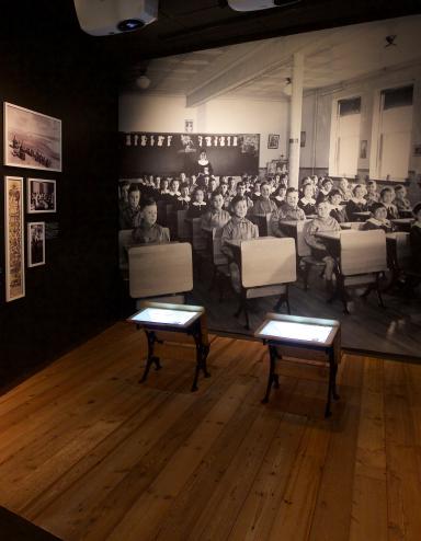 A Museum exhibit showing a black-and-white photo of children sitting in rows at school desks. Two desks, similar to those in the photo sit in the centre of the exhibit. A headline on a text panel reads “Childhood Denied.”