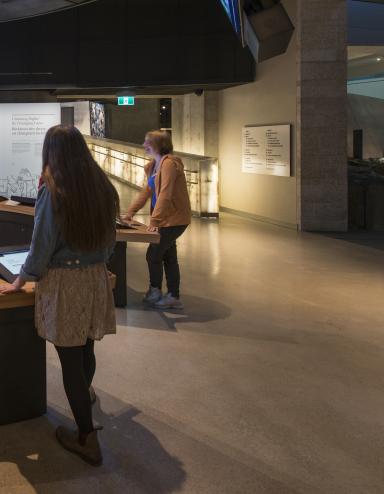 Six people in a museum gallery stand around a circular counter with video screens both embedded in the counter and hanging above them. Partially obscured.