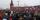 A crowd of people marches holding the red, white and blue striped flags of Russia. Behind is a red concrete wall that ends in a spire.