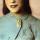 A head-and-shoulder portrait of a smiling Viola Desmond. She is wearing a light blue jacket with embroidered patterns on it. The jacket is held together at the collar by a large pin in the shape of a hand making a “V for victory” symbol.