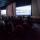 A seated audience viewing three large screens in a dimly lit room.