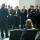 A choir of men wearing black outfits hold books and sing in front of a conductor.