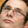 Close-up of a man’s face looking skyward in a thoughtful pose. He is wearing glasses and there are blurry spots of light in the background.