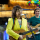 Two guitar players in bright and colourful outfits sing into microphones in front of a balloon arch.