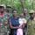 A woman is holding a baby, while four men in army fatigues stand beside and behind her. They are all standing in front of a forest, posed for the camera.