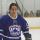Un jeune homme portant un uniforme de hockey bleu orné d'un logo de couronne sur une patinoire intérieure.