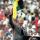 Nelson Mandela, an older dark-skinned man with short grey hair, stands in front of a podium and speaks into two microphones. He is wearing a grey suit with a tie. He is raising his right arm above his head and his hand is making a fist.