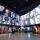 People explore a Museum gallery with two rows of square portraits above a series of alcoves, containing photographs, videos, and text. The alcove on the left contains suspended red dresses.