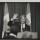 A black and white photograph of Nelson Mandela standing in front of a podium. He is smiling and waving.