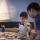 A young child interacting with photos on a table. An adult sits beside him.