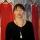 Jaime Black stands in front of red dresses suspended in air on hangers in front of a backdrop. The backdrop features an image of a birch wood forest with more red dresses hanging in it.