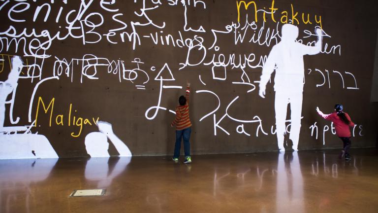 A video of people’s silhouettes is projected onto a large wall. They are writing the word “welcome” in many languages. Two children interact with the video. Partially obscured.