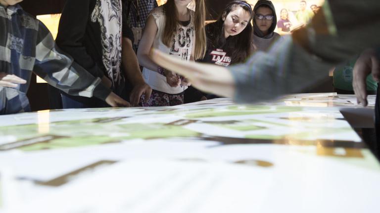 A group of students look at the top of an interactive table emitting white light. Partially obscured.