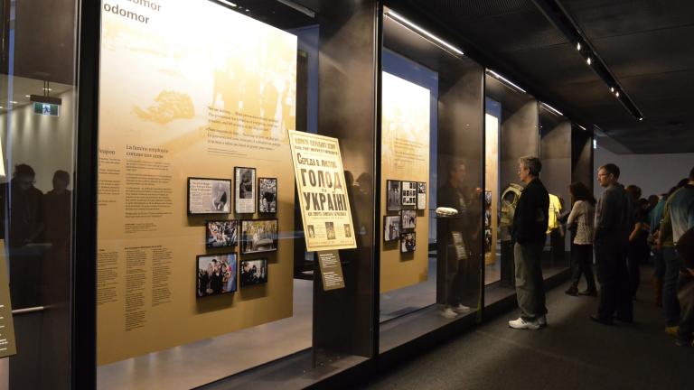 A group of people look at a series of floor-to-ceiling glass display cases. The cases contain large text panels and smaller panels that contain images. Partially obscured.
