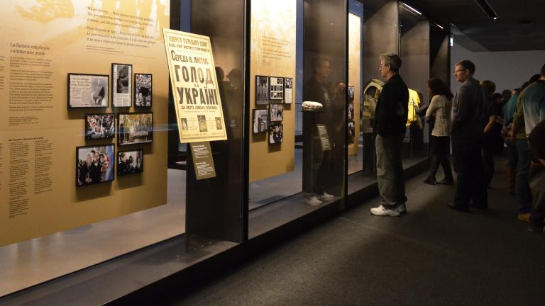 A group of people look at a series of floor-to-ceiling glass display cases. The cases contain large text panels and smaller panels that contain images. Partially obscured.