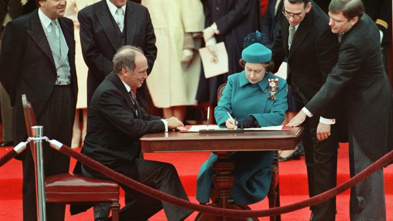 Queen Elizabeth II sitting at a table and signing a document. A man is also sitting to her right, and others are standing behind them around the table.