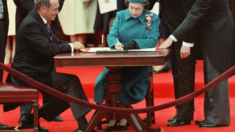 Queen Elizabeth II sitting at a table and signing a document. A man is also sitting to her right, and others are standing behind them around the table.