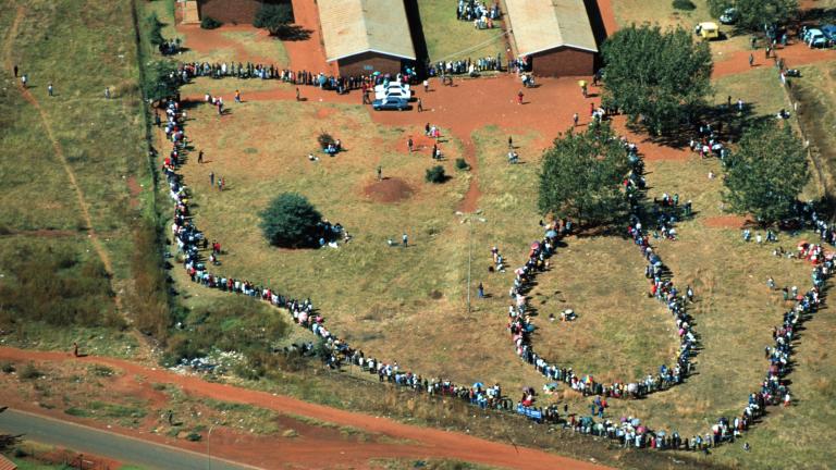  View from above of people standing outside in lines. The ground is mostly covered in grass and the exposed earth is of a reddish colour.