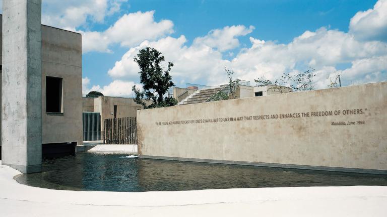 A view of the Apartheid Museum. In the foreground is a pool of water. Rising out of the pool is a large wall with a quote from Nelson Mandela written on it. It reads: “To be free is not merely to cast off one’s chains, but to live in a way that respects and enhances the freedom of others.”