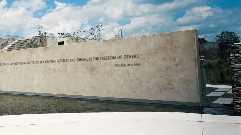 A view of the Apartheid Museum. In the foreground is a pool of water. Rising out of the pool is a large wall with a quote from Nelson Mandela written on it. It reads: “To be free is not merely to cast off one’s chains, but to live in a way that respects and enhances the freedom of others.”