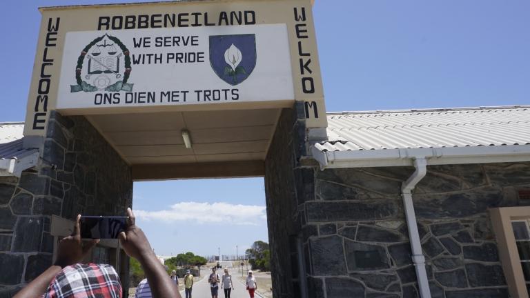 A opening through a building with a large sign over it. The sign reads “Welcome” and “We serve with pride” in both English and Afrikaans. At the top of the sign it says “Robbeneiland.”