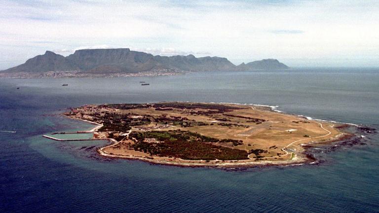An aerial view of Robben Island. The island is very flat and roughly oval in shape, with a little under half its surface covered with trees. An airfield is visible on the right side of the island. In the distant background behind the island, the mainland can be seen, including the buildings of a city. Rising above the city are large mountains. 